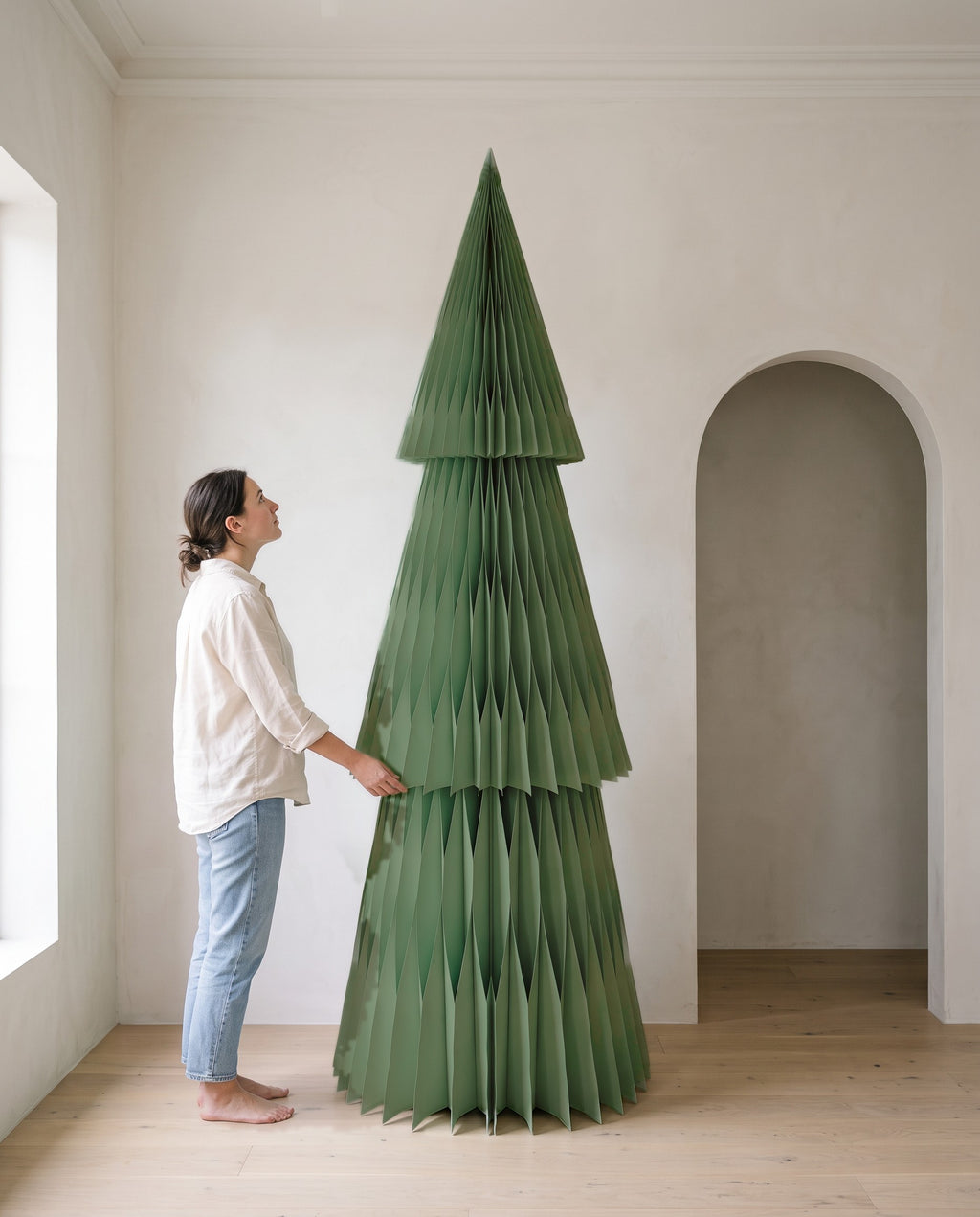 A woman standing beside a 240cm Forma paper Christmas tree in Sage, showing its height